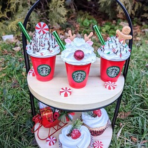 May include: A two-tiered wooden serving tray with a black metal stand. The top tier has three red and white Starbucks cups with whipped cream and holiday decorations. The bottom tier has two cupcakes with white frosting and red berries. The tray is sitting on green grass.