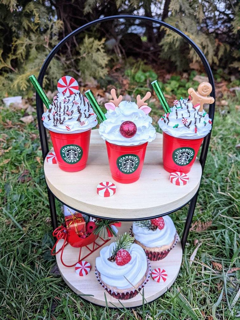 May include: A two-tiered wooden serving tray with a black metal stand. The top tier has three red and white Starbucks cups with whipped cream and holiday decorations. The bottom tier has two cupcakes with white frosting and red berries. The tray is sitting on green grass.