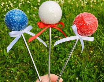 May include: Three cake pops decorated with white, blue, and red sprinkles. Each cake pop has a white ribbon tied around the stick.