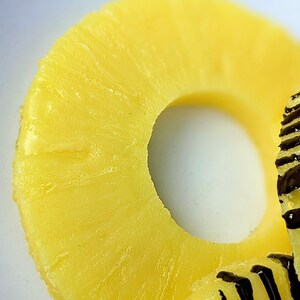 May include: A close-up of a yellow pineapple ring with a hole in the center. The ring is on a white surface.