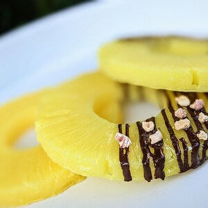 May include: Three pineapple rings on a white plate. The top ring is decorated with chocolate stripes and pink sprinkles.