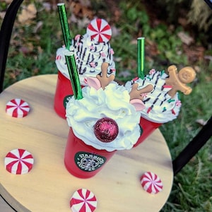 May include: Three red cups with whipped cream, candy canes, and a reindeer and gingerbread man topper. The cups are on a wooden tray with red and white candy canes. The cups have the Starbucks logo on them.