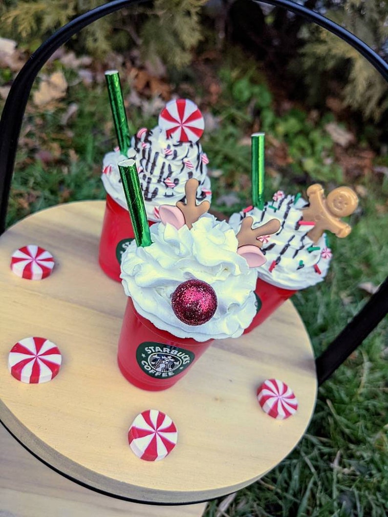 May include: Three red cups with whipped cream, candy canes, and a reindeer and gingerbread man topper. The cups are on a wooden tray with red and white candy canes. The cups have the Starbucks logo on them.