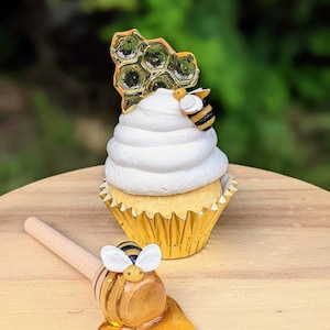 Puede incluir: Un cupcake blanco con glaseado amarillo cubierto con un panal y una pequeña figura de abeja. Un cucharón de miel de madera con una figura de abeja y miel goteando de él se encuentra sobre una superficie de madera.