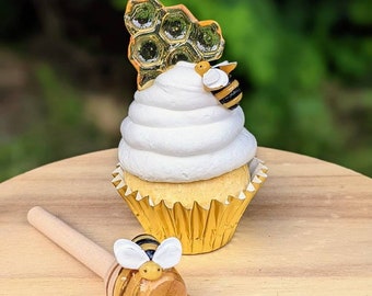 May include: A white cupcake with yellow frosting topped with a honeycomb and a small bee figurine. A wooden honey dipper with a bee figurine and honey dripping from it sits on a wooden surface.