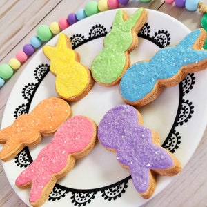 May include: Six pastel-colored bunny-shaped sugar cookies decorated with sprinkles on a white plate with a black lace design around the edge.