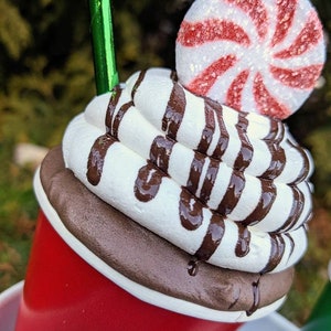 Op de afbeelding: Een decoratieve rode beker met een witte slagroomtopping en chocoladesiroop drizzle. Een pepermunt snoepje ligt erop. De beker heeft een groen rietje en een logo met het woord "Merry".