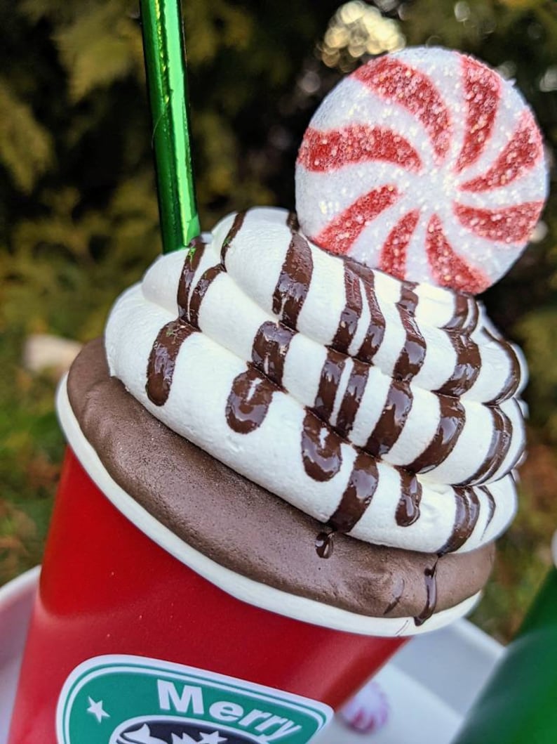 Op de afbeelding: Een decoratieve rode beker met een witte slagroomtopping en chocoladesiroop drizzle. Een pepermunt snoepje ligt erop. De beker heeft een groen rietje en een logo met het woord "Merry".
