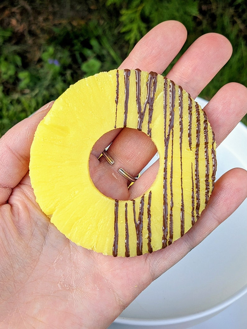 May include: A pineapple ring with chocolate drizzled on top. The ring is held in a hand, with the chocolate side facing the camera.