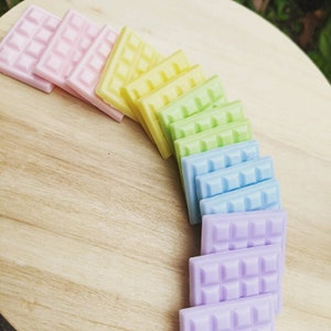 May include: A rainbow of wax melts arranged in a semi-circle on a wooden surface. The wax melts are in shades of pink, yellow, green, blue, and purple.