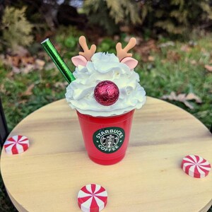 May include: A red Starbucks coffee cup with whipped cream, a red glitter ball, and reindeer antlers. The cup is on a wooden table with red and white peppermint candies.