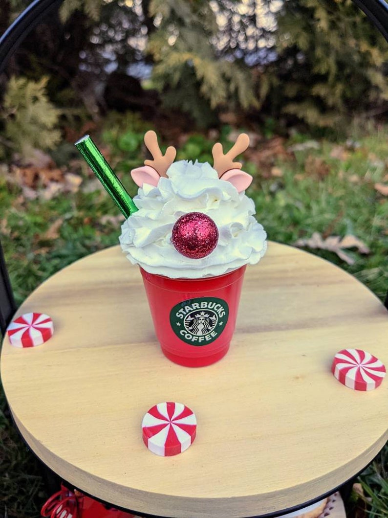 May include: A red Starbucks coffee cup with whipped cream, a red glitter ball, and reindeer antlers. The cup is on a wooden table with red and white peppermint candies.
