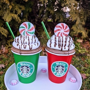 Op de afbeelding: Twee decoratieve bekers met vakantiethema, &eacute;&eacute;n groen en &eacute;&eacute;n rood, met de tekst "Merry & Bright". Elke beker heeft een topping van slagroom met chocoladedrizzle, een pepermunt snoepje en een groen rietje. Pepermunt snoepjes zijn verspreid.