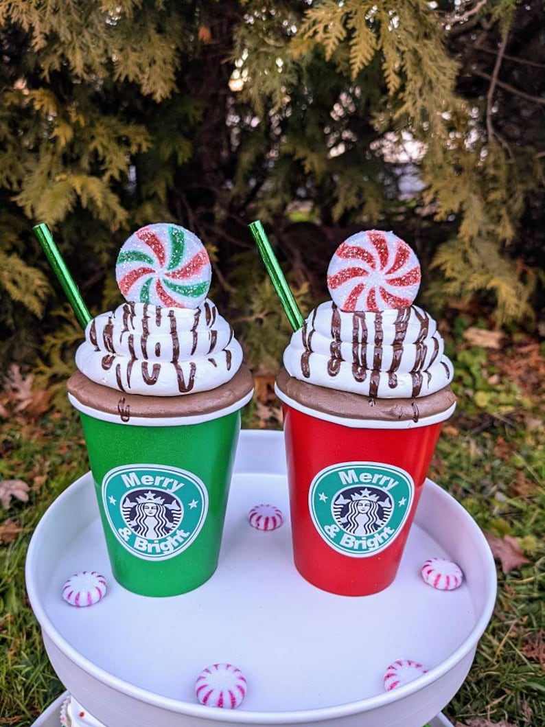 Op de afbeelding: Twee decoratieve bekers met vakantiethema, &eacute;&eacute;n groen en &eacute;&eacute;n rood, met de tekst "Merry & Bright". Elke beker heeft een topping van slagroom met chocoladedrizzle, een pepermunt snoepje en een groen rietje. Pepermunt snoepjes zijn verspreid.