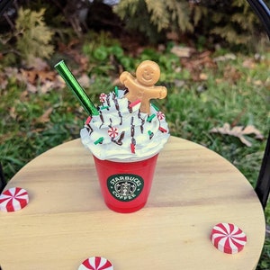 May include: A red Starbucks cup with whipped cream, chocolate drizzle, green sprinkles, and a gingerbread man on top. The cup is sitting on a wooden surface with three red and white peppermint candies.