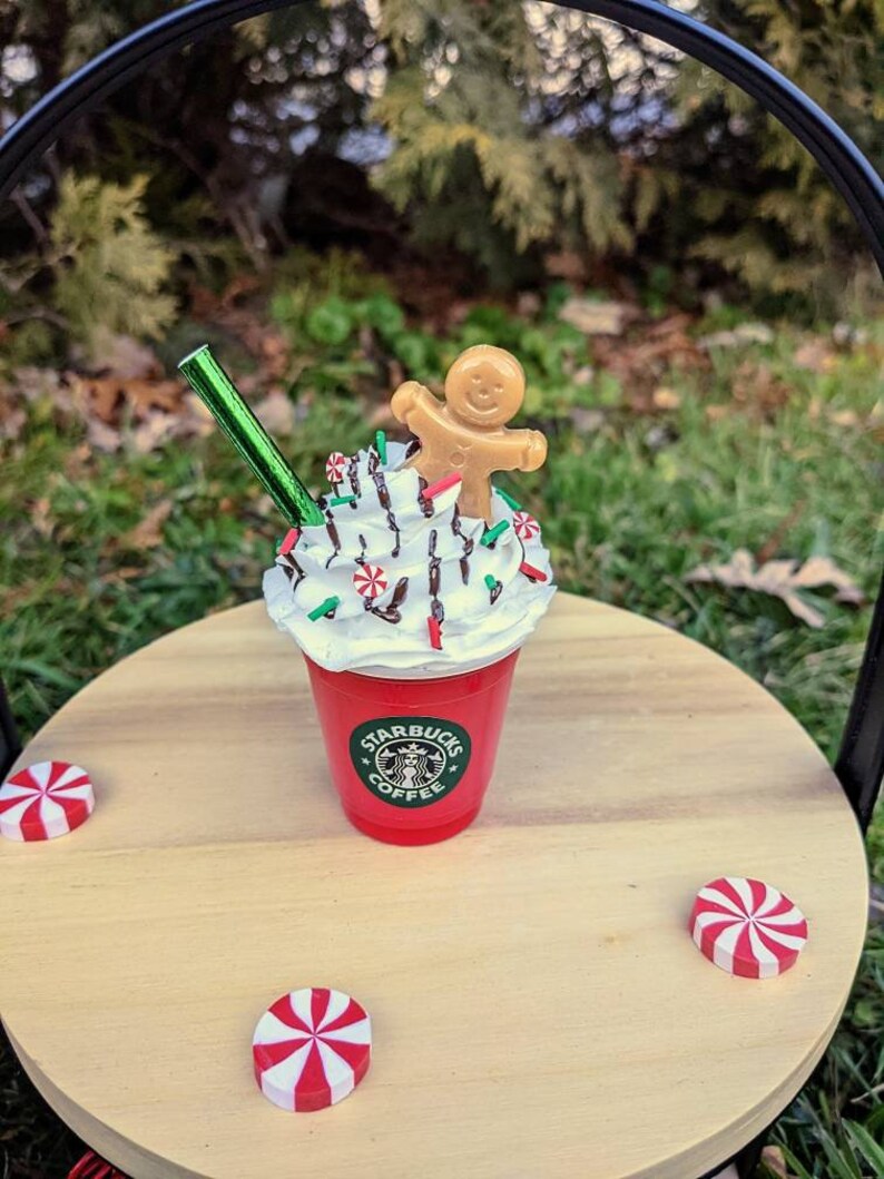 May include: A red Starbucks cup with whipped cream, chocolate drizzle, green sprinkles, and a gingerbread man on top. The cup is sitting on a wooden surface with three red and white peppermint candies.