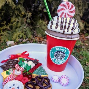 Op de afbeelding: Een feestelijke display met een rode beker met slagroom, chocoladesaus en een pepermunt snoepje. Een dienblad bevat koekjes, snoepjes en pretzels. De beker heeft de tekst "Merry & Bright".