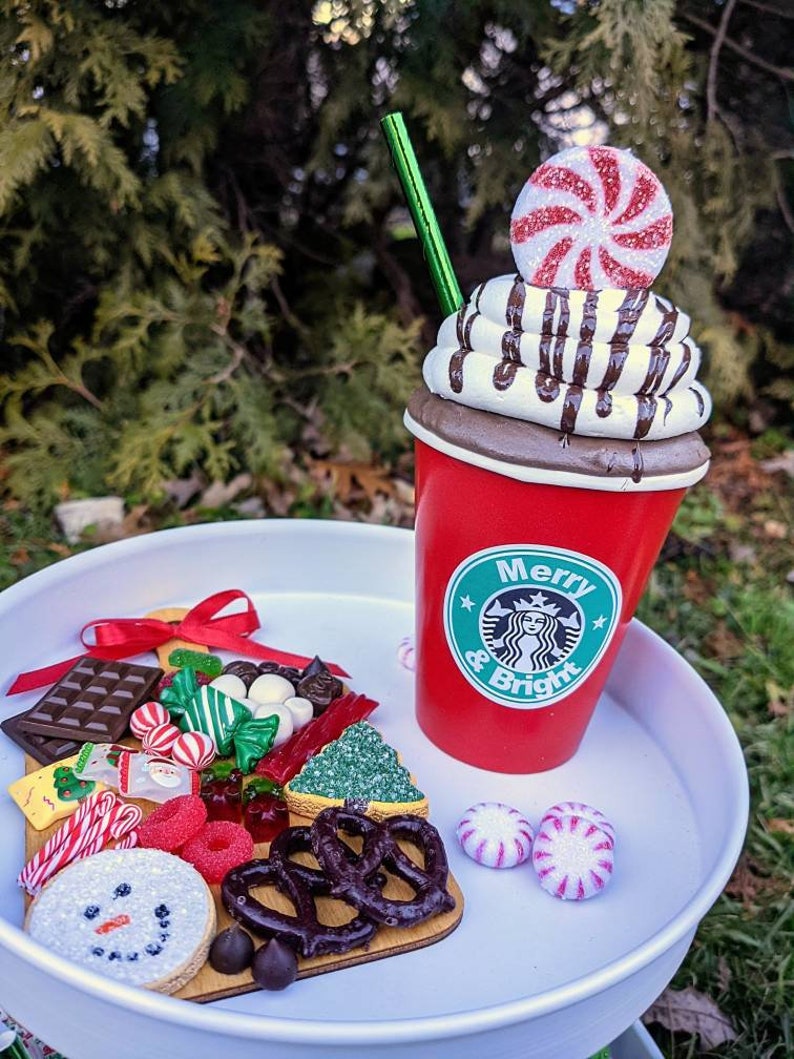 Op de afbeelding: Een feestelijke display met een rode beker met slagroom, chocoladesaus en een pepermunt snoepje. Een dienblad bevat koekjes, snoepjes en pretzels. De beker heeft de tekst "Merry & Bright".