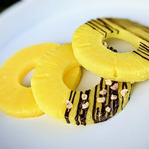 May include: Three pineapple rings on a white plate. Two of the rings are plain, while the top ring has chocolate stripes and sprinkles.