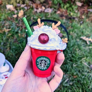 May include: A red Starbucks coffee cup with a white whipped cream top, a red glitter ball, and reindeer antlers. The cup has a green Starbucks logo with the text "Starbucks Coffee".