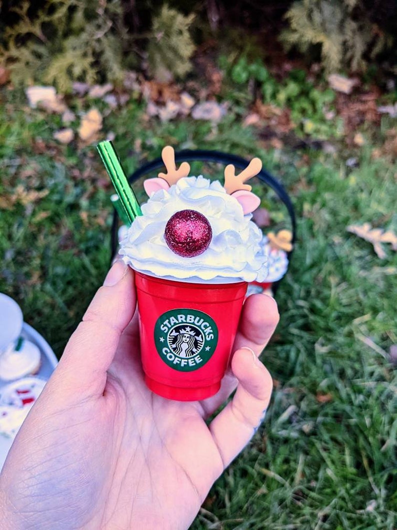 May include: A red Starbucks coffee cup with a white whipped cream top, a red glitter ball, and reindeer antlers. The cup has a green Starbucks logo with the text "Starbucks Coffee".