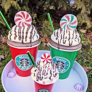Op de afbeelding: Drie feestelijke koffiebekers, rood en groen, versierd met slagroom, chocoladesaus en pepermunt snoepjes. De bekers hebben de tekst "Merry & Bright" en een logo. Een wit dienblad bevat de bekers en snoepjes.