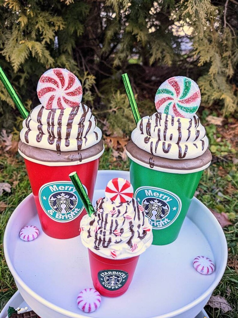 Op de afbeelding: Drie feestelijke koffiebekers, rood en groen, versierd met slagroom, chocoladesaus en pepermunt snoepjes. De bekers hebben de tekst "Merry & Bright" en een logo. Een wit dienblad bevat de bekers en snoepjes.