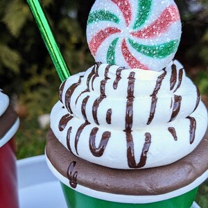 Op de afbeelding: Een feestelijke decoratieve beker die lijkt op warme chocolademelk. De groene beker heeft een witte rand en een bruine basis, gegarneerd met slagroom en chocoladesaus. Een pepermunt snoepje en een groen rietje maken het ontwerp compleet.