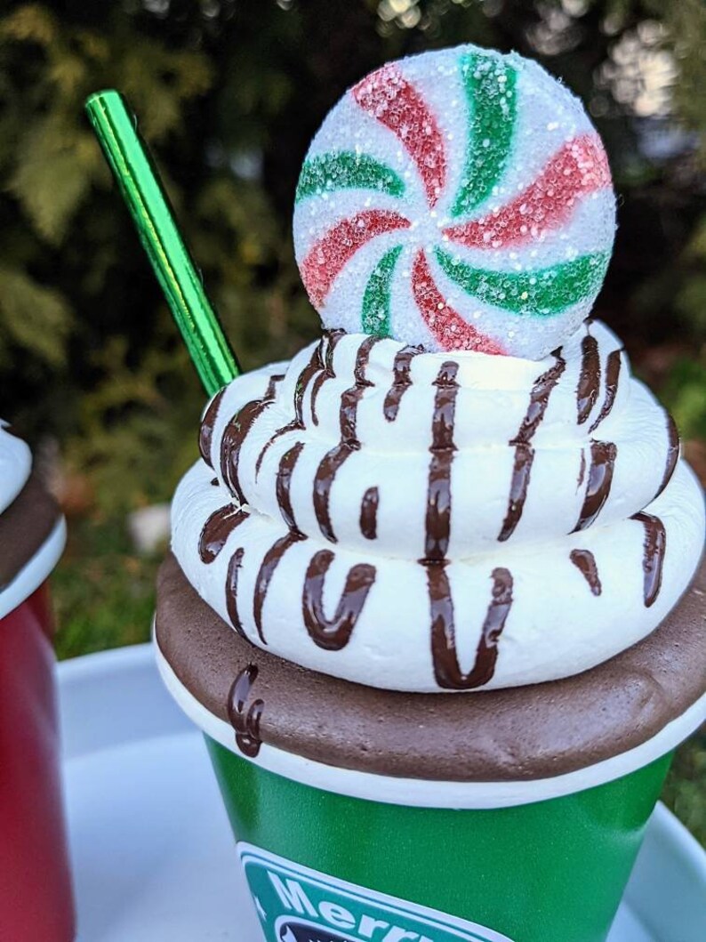 Op de afbeelding: Een feestelijke decoratieve beker die lijkt op warme chocolademelk. De groene beker heeft een witte rand en een bruine basis, gegarneerd met slagroom en chocoladesaus. Een pepermunt snoepje en een groen rietje maken het ontwerp compleet.