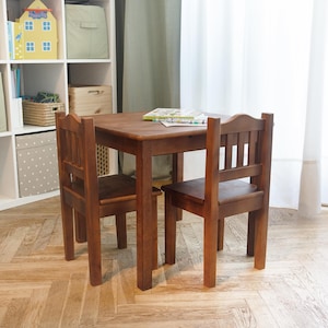 Peut inclure: Un ensemble de table et deux chaises en bois pour enfants. La table est brune et a un plateau carré. Les chaises sont également brunes et ont un dossier à lattes.