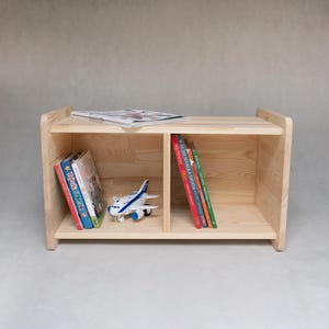Peut inclure: Une étagère en bois avec deux étagères, chacune divisée en deux compartiments. Les étagères sont remplies de livres et d'un avion jouet. L'étagère est en bois naturel et a un design simple.