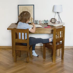 Pode incluir: Uma criança está sentada numa pequena secretária de madeira com uma cadeira a condizer, a escrever num caderno. A secretária tem uma lâmpada, livros e letras decorativas que soletram "KELOWS". A sala tem chão de parquet e um quadro emoldurado na parede.