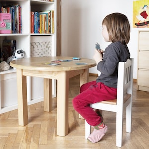 Op de afbeelding: Een ronde tafel van lichtgekleurd hout met vier stevige poten, gecombineerd met een bijpassende houten stoel met een wit frame en een zitting van natuurlijk hout. Het tafelblad is glad en rond en biedt een stabiel oppervlak.