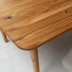 Peut inclure: Gros plan sur une table en bois avec une finition naturelle. La table a un plateau rectangulaire aux coins arrondis et quatre pieds effilés. Le grain du bois est visible, et la table semble être en bois massif. La table est de couleur marron clair.