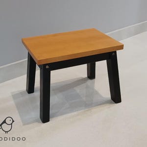 May include: A small wooden stool with a rectangular, light brown seat and black legs. The stool is a simple design, suitable for various uses. The legs are square and sturdy, providing stability. The brand logo "WOODIDOO" is visible in the bottom left corner.