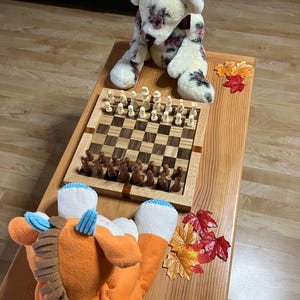 May include: A wooden chess set with brown and white pieces is displayed on a wooden table. A plush bear with floral patterns and an orange and blue plush toy are also on the table. Decorative fall leaves are scattered around.