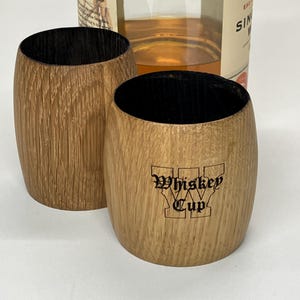 May include: Two barrel-shaped wooden cups with dark interiors and natural wood grain exteriors. One cup features the black text Whiskey Cup printed on the front. A glass bottle containing amber liquid is positioned behind the two wooden cups.
