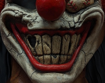 Puede incluir: Primer plano de una máscara de payaso aterradora. La máscara presenta una gran nariz roja, una amplia sonrisa agrietada con dientes podridos y una pelota de béisbol cosida en la cuenca del ojo. Cabello rojo visible a los lados.