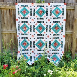 Op de afbeelding: Een wit quilt met een herhalend patroon van turquoise, zwarte en roze vierkanten. Het quilt hangt aan een houten hek.