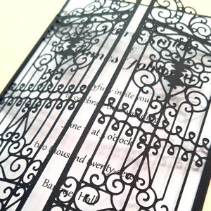 May include: A wedding invitation featuring a black laser-cut paper overlay with an intricate gate design. The overlay sits atop a white card with black printed text that reads, "joyfully invite you to celebrate the wedding," "at 5 o'clock," and "two thousand twenty-seven."