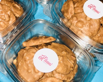 Peut inclure: Gros plan sur un récipient en plastique transparent rempli de pralines aux noix de pécan. Les pralines sont de couleur brun clair et ont une forme légèrement irrégulière. Un autocollant blanc avec le texte "P.Leauns Fine Candies" est visible sur le récipient.