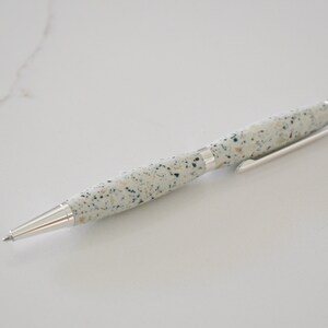 May include: A white pen with a speckled pattern and silver accents. The pen is lying on a white surface.
