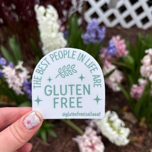 Puede incluir: Una pegatina blanca con un dise&ntilde;o verde claro y el texto "THE BEST PEOPLE IN LIFE ARE GLUTEN FREE" con el nombre de usuario de Instagram @glutenfreeisallaeat. La pegatina se sostiene frente a un fondo de flores.