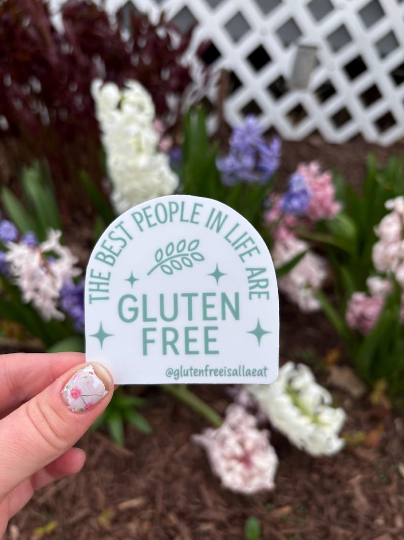 Puede incluir: Una pegatina blanca con un dise&ntilde;o verde claro y el texto "THE BEST PEOPLE IN LIFE ARE GLUTEN FREE" con el nombre de usuario de Instagram @glutenfreeisallaeat. La pegatina se sostiene frente a un fondo de flores.