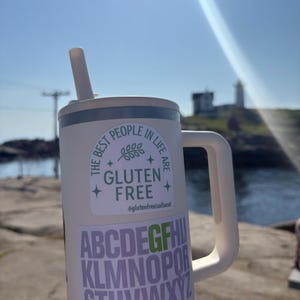 Puede incluir: Un vaso aislado blanco con asa y pajita. El vaso tiene dos pegatinas: una con el texto "THE BEST PEOPLE IN LIFE ARE GLUTEN FREE" y otra con el alfabeto. El fondo muestra un d&iacute;a soleado con un faro.