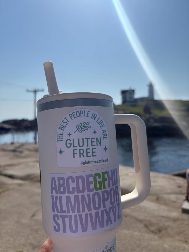 Puede incluir: Un vaso aislado blanco con asa y pajita. El vaso tiene dos pegatinas: una con el texto "THE BEST PEOPLE IN LIFE ARE GLUTEN FREE" y otra con el alfabeto. El fondo muestra un d&iacute;a soleado con un faro.