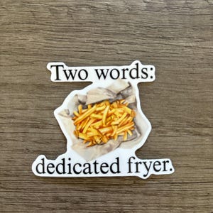 May include: A white sticker with the text "Two words: dedicated fryer." The sticker features a graphic of golden french fries in a paper container. The sticker is on a brown wooden surface.