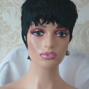 Peut inclure: Tête de mannequin avec une perruque noire courte coiffée en chignon. Le mannequin est entièrement maquillé, avec du rouge à lèvres rose et de l'ombre à paupières pailletée argentée. Boucles d'oreilles dorées visibles.