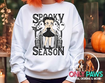 May include: White sweatshirt with a graphic featuring Betty Boop in a black dress, a ghost, and the words "Spooky Season." The design includes bats, skulls, and a decorative frame. The sweatshirt is part of a Halloween-themed collection.