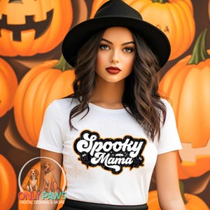 以下が含まれることがあります： 黒い帽子と「Spooky Mama」の文字が白黒でデザインされた白いTシャツを着た女性。背景にはオレンジ色のカボチャが描かれています。Tシャツのデザインはオレンジ色の縁取りがあります。